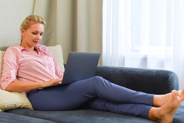 blonde woman 30 years old works on a computer at home on the couch.