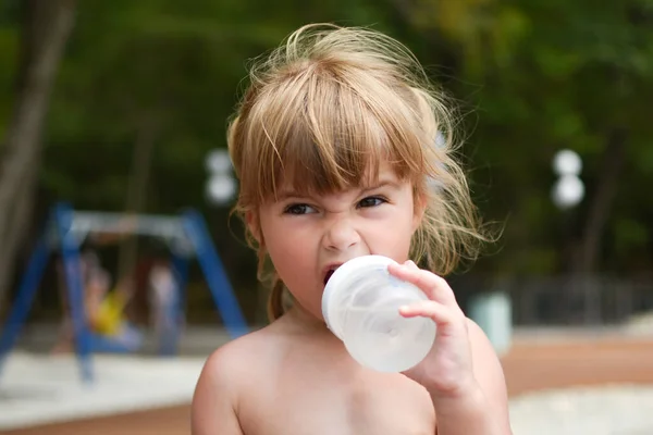 Забавная маленькая девочка с бутылкой выпивает на открытом воздухе. Веселый портрет девочки 3 года. — стоковое фото