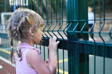 Little child girl looks behind a metal grate fence. A girl in an orphanage clipart