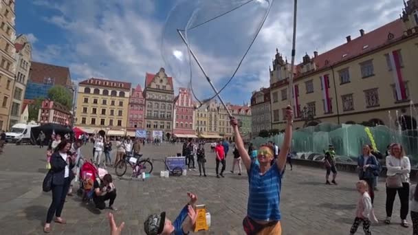 Человек запускает огромные мыльные пузыри, развлекая людей в старом центре города. Широкий угол, медленная съемка . — стоковое видео