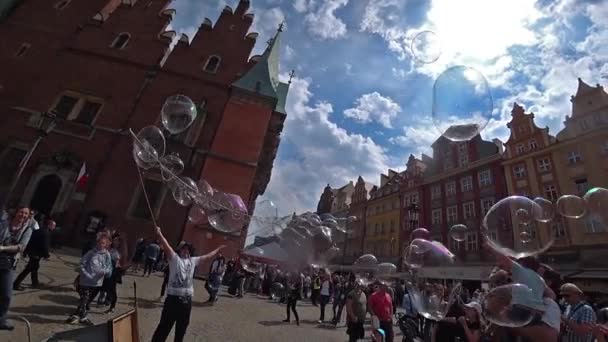 Человек запускает большие мыльные пузыри, развлекая людей в старом центре города. Широкий угол, медленная съемка . — стоковое видео