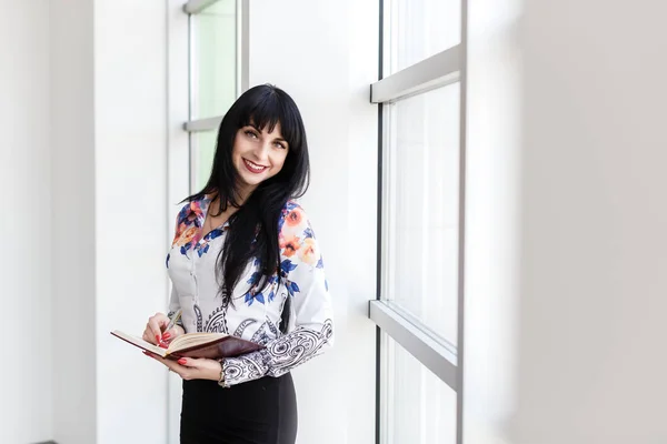 Junge glückliche Frau, die am Fenster steht und in ein Notizbuch schreibt — Stockfoto