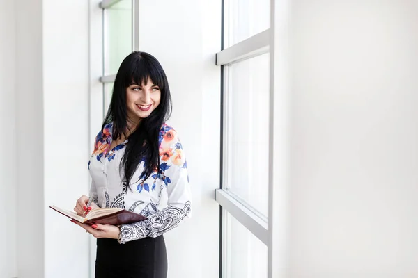 Joven Hermosa mujer feliz de pie cerca de la ventana, escribiendo en un n — Foto de Stock