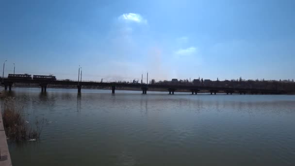 Silueta del tranvía que monta en el puente sobre el río sobre un fondo de fábrica, retroiluminado . — Vídeos de Stock