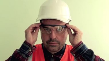 Yansıtıcı yelek ve beyaz kask giymiş kafkas traşsız adam işçi koruyucu çalışma gözlükleri ve kulak tıkacı koyar. Açık yeşil arka planda izole... Endüstriyel güvenlik konsepti.
