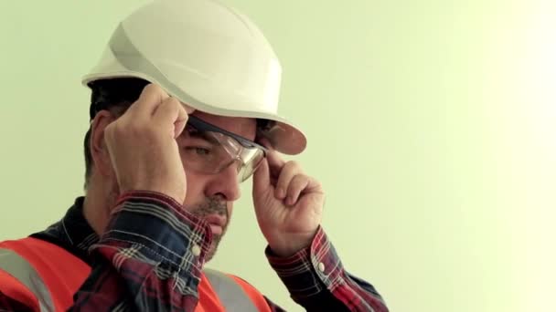 反射ベストと白いヘルメットに身を包んだ白人の剃毛されていない男性労働者は 薄緑色の背景に隔離された保護作業眼鏡を着用します 産業安全コンセプト — ストック動画
