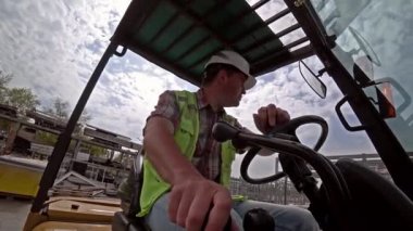 Kafkas adam, yansıtıcı yelek ve beyaz kask giymiş bir forklift açık havada çalışan depo işçisi. Aksiyon kamerası çekimi, geniş açı.