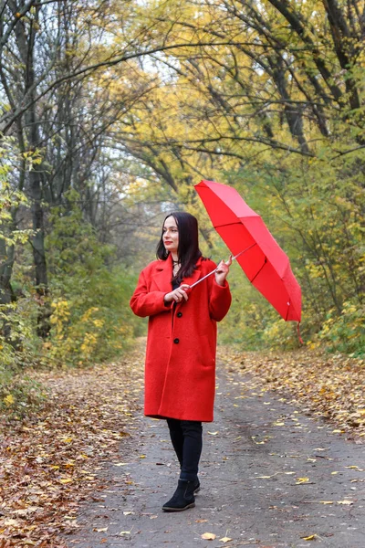 Beautiful woman dressed in a red coat walk in a city park with a red umbrella at autumn day.