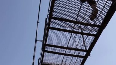 İnsanlar sallanan metal kafes asma köprüde yürüyorlar. Mavi gökyüzüne karşı delikli zemin den aşağıdan görünümü.