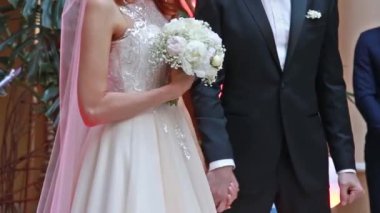 Gelin ve damat bir düğün törenine el tutuyorlar. Beyaz elbise, gül buket tutan kadın.