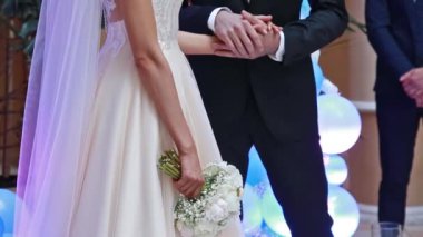 Gelin ve damat bir düğün törenine el tutuyorlar. Beyaz elbise, gül buket tutan kadın.