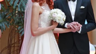 Gelin ve damat bir düğün törenine el tutuyorlar. Beyaz elbise, gül buket tutan kadın.