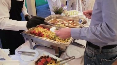 Siyah eldivenli garson, şenlikli bir resepsiyonda misafirlerin tabaklarına yemek koyar..