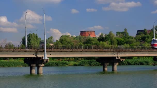 Трамвай рухається вздовж мосту над річкою на тлі рослини і блакитного хмарного неба . — стокове відео