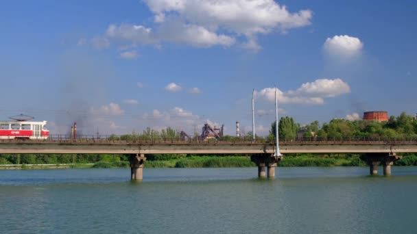 Tram si muove lungo il ponte sul fiume sullo sfondo di piante e cielo nuvoloso blu . — Video Stock