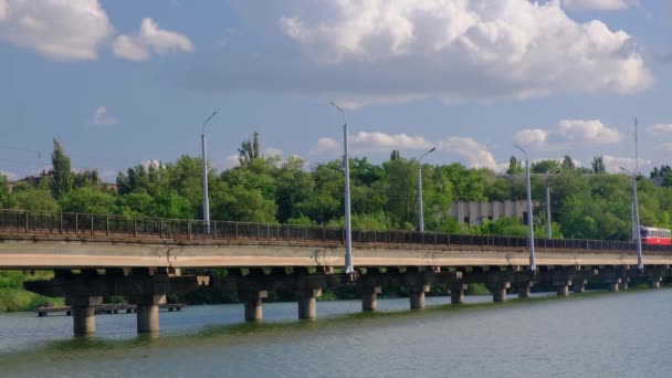 Трамвай движется по мосту через реку в городе на фоне зеленых деревьев . — стоковое видео