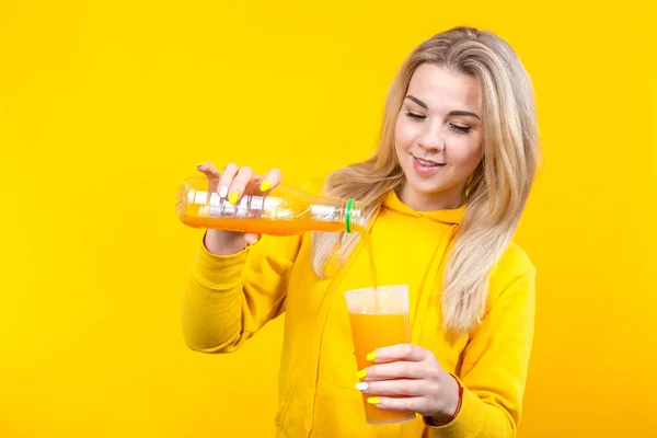 魅力的な若いですブロンド女性でカジュアルスポーティーな服を注ぐオレンジジュースからプラスチックボトルへガラス 笑顔の女の子は黄色の背景に隔離 — ストック写真