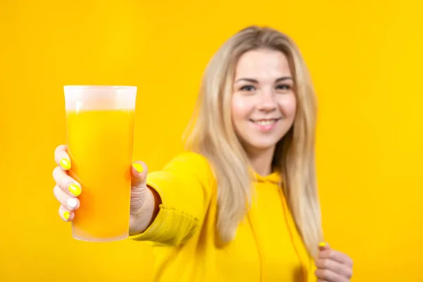 穿着黄色休闲装 拿着橙汁杯的快乐而美丽的金发女青年 带着黄色背景的微笑女孩 看着相机 — 图库照片