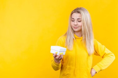 Sarı spor takım elbiseli, sarı arka planda izole edilmiş, gülümseyen, kutuya bakan beyaz bir hediye kutusu olan beyaz, güzel, beyaz bir sarışın kadının portresi..