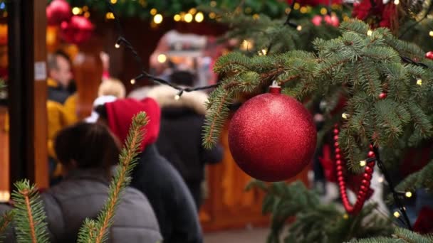 在一个城市的集市上 一个红色的圣诞舞会挂在一棵天然的圣诞树上 一群人在一个喜庆的广场上散步 — 图库视频影像