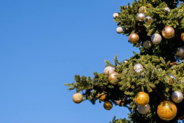 Güneşli yaz gününde mavi bir arka planda sahte Noel ağacının dalları üzerinde altın ve beyaz yeni yıl süslemeleri ve çelenkler. İnsan yok, kar yok. Boşluğu kopyala.