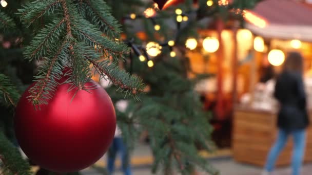 市内のフェアで自然の新年の木にぶら下がっている赤いクリスマスボールの閉鎖 お祝いの広場を歩いている人々の青白い群衆 — ストック動画