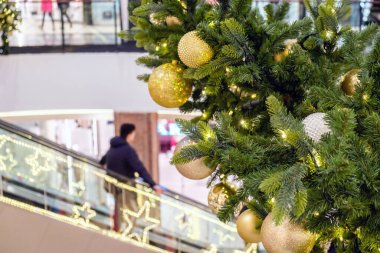 Altın Noel topları ve süslenmiş bir köknar ağacının üzerindeki çelenk Alışveriş Merkezi 'nde. Bulanık insanlar yürüyen merdivende, şenlikli iş merkezinde yeni yıl alışverişi yapıyorlar.,.