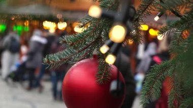 Şehir panayırındaki doğal bir yılbaşı ağacında asılı duran kırmızı bir Noel balosunun yakın çekimi. Bulanık kalabalık bir meydanda yürüyor..