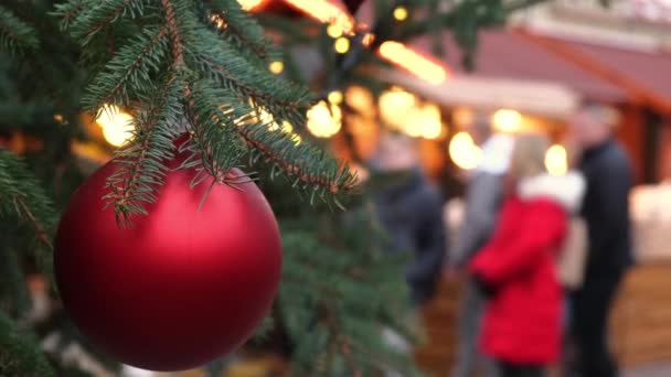 在市集上 一个挂在一棵天然新年树上的红色圣诞舞会的布景 在节日广场上散步的人群模糊不清 — 图库视频影像