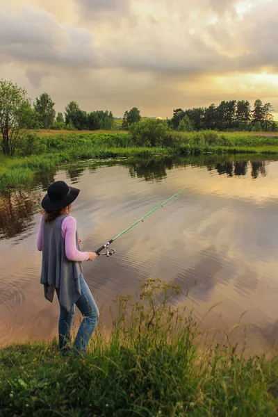 Menina em uma vara de pesca chapéu no lago, paisagem de verão, nuvens de tempestade no céu. atividades ao ar livre. vista traseira . — Fotografia de Stock