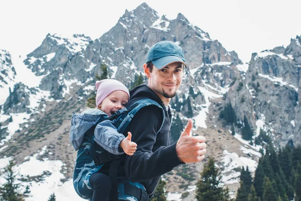 Aantrekkelijke jonge vader met haar baby dochter in ergonomische draagzak op rug buiten in berg natuur, Toon duim omhoog. — Stockfoto