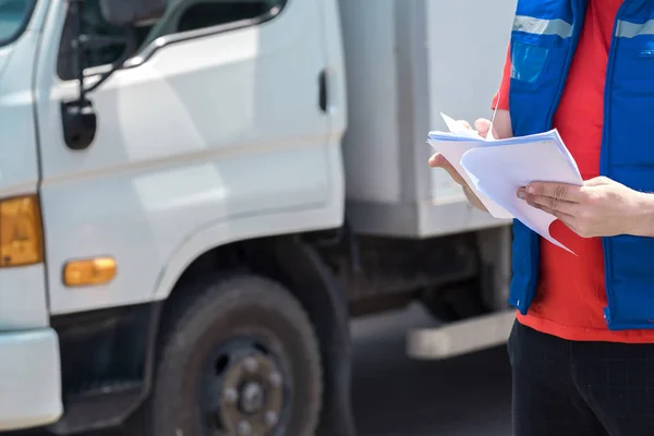 Courier driver in uniform making notes in document and delivery white truck behinde him