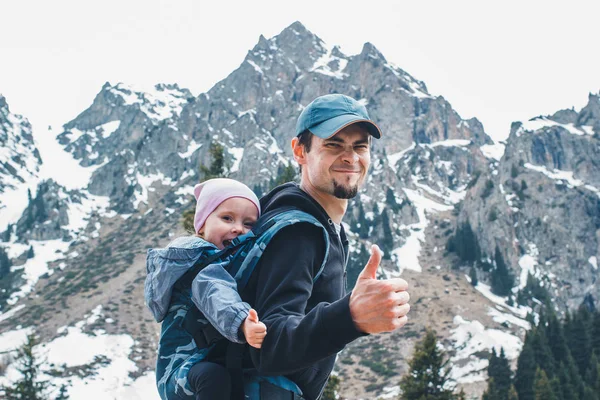 Aantrekkelijke jonge vader met haar baby dochter in ergonomische draagzak op rug buiten in berg natuur, Toon duim omhoog. — Stockfoto