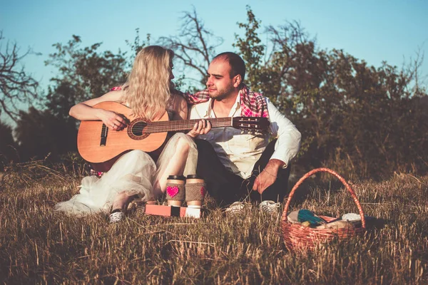 Romantik çift gün batımı ışığında piknik yaparken sarılıyor. Beyaz gömlekli adam ve gitar çalan çekici genç kadın açık havada çimenlerin üzerinde birlikte oturuyorlar.. — Stok fotoğraf