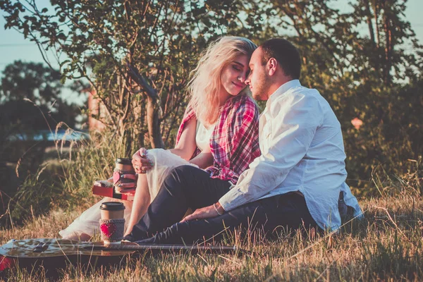 Romantik çift gün batımı ışığında piknik yaparken sarılıyor. Beyaz gömlekli adam ve çekici genç kadın açık havada çim üzerinde birlikte oturuyor. — Stok fotoğraf