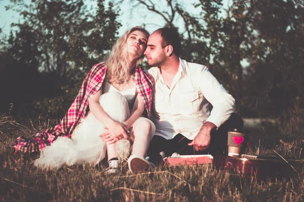 Casal romântico abraçando enquanto faz piquenique à luz do pôr do sol. Homem de camisa branca e atraente jovem mulher estão sentados juntos na grama ao ar livre . — Fotografia de Stock