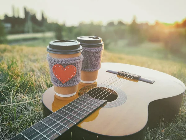 Two paper coffee cup in knitted cozy sleeves standing on guitar. Outdoor picnic sunset scene. — Stock Photo, Image