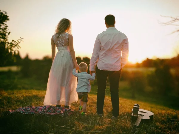 Kleine familie romantisch picknick scène buiten in zonsondergang. Ouders houden handen van kleine peuter jongen tussen hen. — Stockfoto