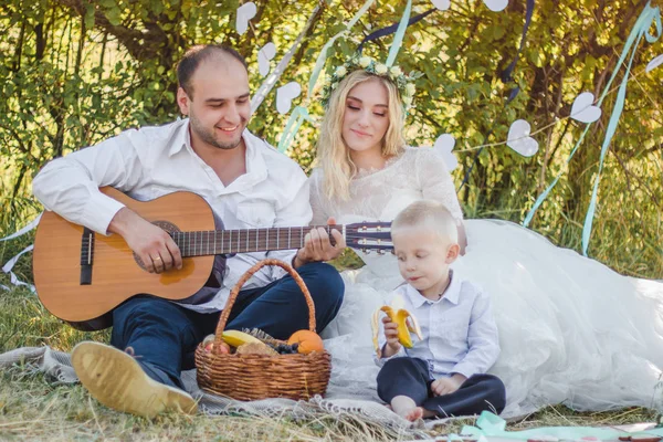 Picknick stijl buiten bruiloft. Bruidegom spelen op gitaar, jonge aantrekkelijke blomde bruid en kleine blonde jongen zittend op een deken in de zomer zonnige dag. — Stockfoto