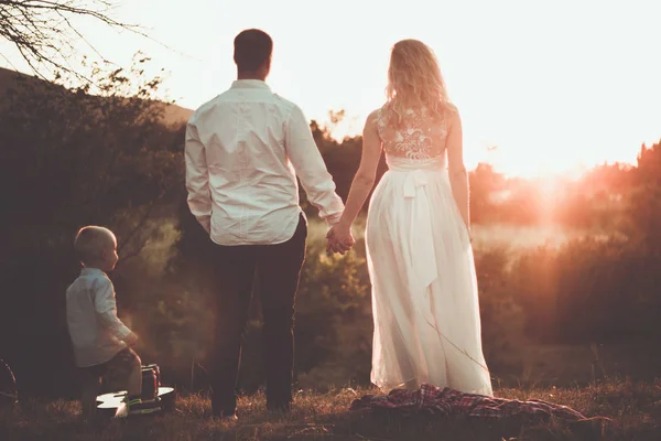 Kleine familie romantisch picknick scène buiten in zonsondergang. Ouders houden van handen en kleine peuter jongen spelen in de buurt van hen. — Stockfoto