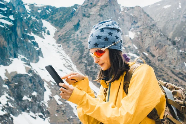 Beautiful young woman hold in hands mobile phone, person type message on smartphone, relax tourist travels planning trip in rock snow mountain, hipster enjoy winter nature
