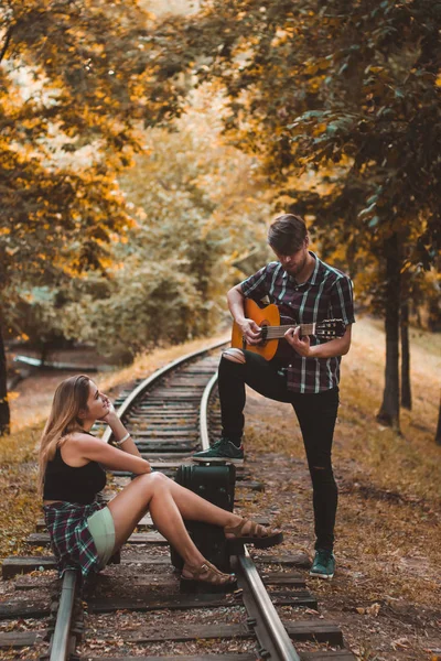 A young couple of lovers missed the train. Playing song with guitar on the rails in the autumn forest waiting for the next train. — Stock Photo, Image