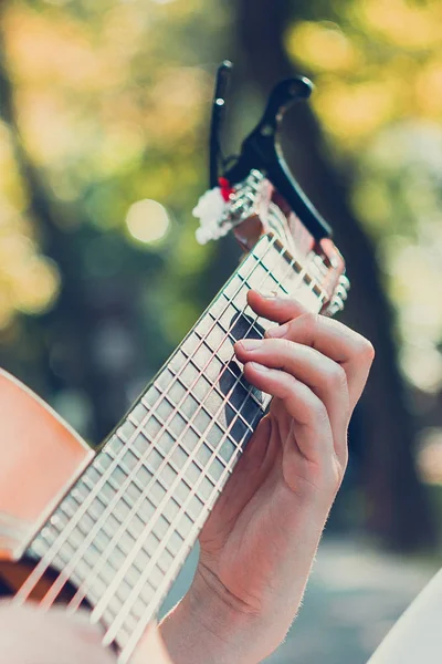 Close up guitar neck with capo in the park. Young man sitting on the bench in the park playing the guitar. Young attractive man enjoys live music in last sunny days autumn holiday. Retro lens used. — Stock Photo, Image