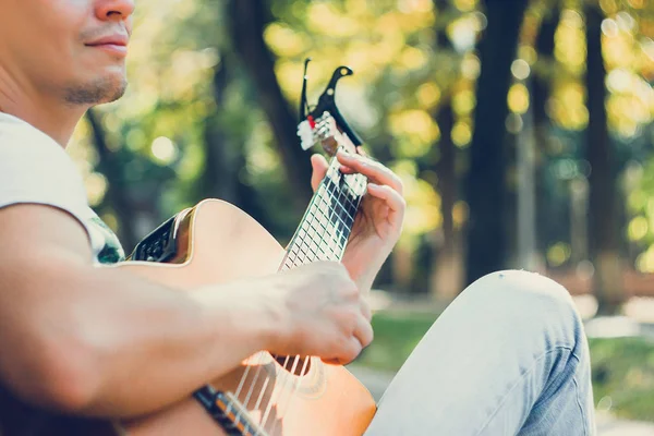 Close up guitar neck with capo in the park. Young man sitting on the bench in the park playing the guitar. Young attractive man enjoys live music in last sunny days autumn holiday. Retro lens used. — Stock Photo, Image