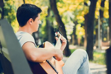 Parktaki bankta oturan genç adam Capo ile akustik gitar çalıyor. Genç ve çekici bir adam son bahar tatilinde güneşli günlerde canlı müzik dinlemekten hoşlanıyor. Retro lens kullanılmış. Seçici yumuşak odak.