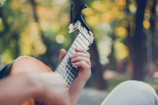 Close up guitar neck with capo in the park. Young man sitting on the bench in the park playing the guitar. Young man enjoys live music in last sunny days autumn holiday. Selective soft focus. — Stock Photo, Image