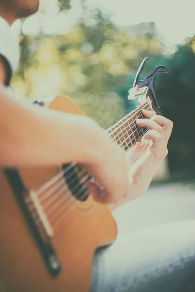 Close up guitar neck with capo in the park. Young man sitting on the bench in the park playing the guitar. Young man enjoys live music in last sunny days autumn holiday. Selective soft focus. — Stock Photo, Image