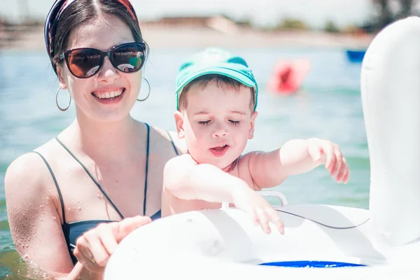 A cute little boy in a blue baseball hat and young attractive mom in sea with an inflatable ring.