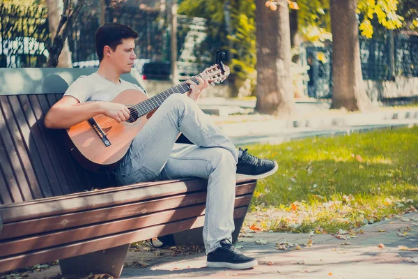 Young man sitting on the bench in the park playing on acoustic guitar with capo. Young attractive man enjoys live music in last sunny days autumn holiday. Retro lens used. Selective soft focus. — Stock Photo, Image