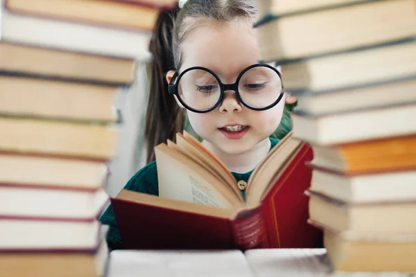 Preschool age girl in big glasses reading a red book behind arch made of books.